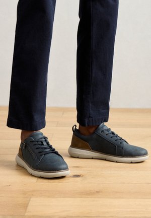 Zapatillas deportivas de cuero azul con cordones grises y un detalle marrón en el talón. Superficie texturizada, suela de goma y un pequeño detalle de cremallera en el lateral.