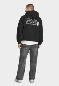 Mężczyzna w czarnej bluzie z kapturem z białym nadrukiem tekstu i grafik na plecach, szare dżinsy, białe trampki, stoi tyłem na białym tle.