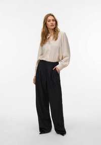 Kreemikas nööpidega särgi tekstuurse kangaga, kombineeritud kõrge vöökohaga musta laiade pükstega. Modellil on käed taskutes ja soeng on stiilselt vormistatud.