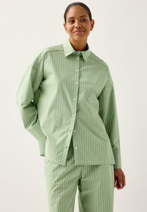 Kvinde med bundet hår, iført løs, lysegrøn, hvidstribet skjorte med knapper og matchende bukser, der smiler let mod en ensfarvet baggrund.