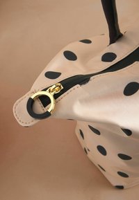 Beige kabelka s bodkami a čiernym zipsom, zlatými kovovými detailmi a čiernymi akcentmi. Hladký povrch s guľatým tvarom a pevným držadlom.