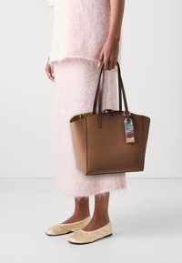 Brun læder tote taske med to håndtag, et kontrastfyldt gult indre og et multicolor mærkat. Båret med et pink fuzzy outfit og flade sko.
