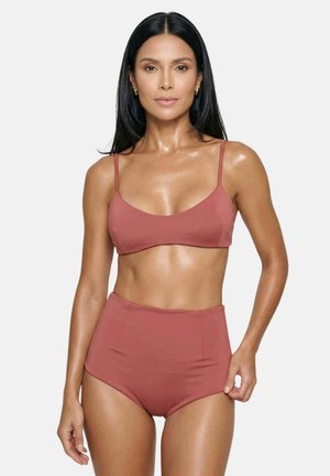 Frau mit langen dunklen Haaren, die einen hochgeschnittenen Bikinihöschen in Rostfarbe und ein passendes Bralette trägt, steht vor einem schlichten hellen Hintergrund.