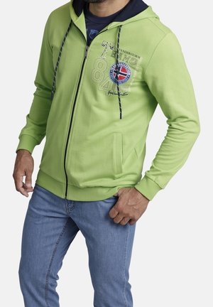 Man wearing lime green zip-up hoodie with blue interior hood, graphic text, and light blue jeans, standing with one hand in pocket.