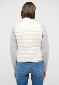 Elfenbeinfarbene wattierte Weste mit hohem Kragen, glatter Textur und horizontalen Steppnähten. Kombiniert mit einem hellgrauen Pullover und blauen Jeans.