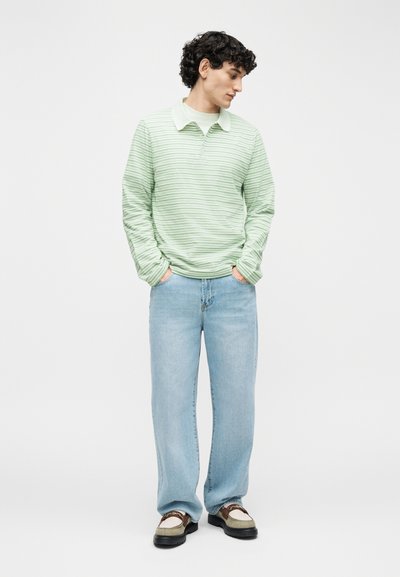 Άνδρας με σγουρά μαλλιά που φοράει ανοιχτό πράσινο πουκάμισο πόλο με μακριά μανίκια ριγέ, ανοιχτό μπλε τζιν και μπεζ και καφέ μοκασίνια, στέκεται με τα χέρια στις τσέπες.