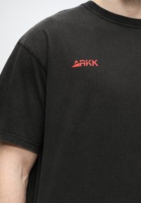 Μαύρο κοντομάνικο μπλουζάκι με κόκκινο λογότυπο "ARKK" εκτυπωμένο στο πάνω μέρος του στήθους, φορεμένο από ένα άτομο με μερικώς ορατό χέρι και λαιμό.