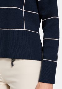 Marineblauer Pullover mit cremefarbenem Gittermuster, gerippten Bündchen und seitlichem Schlitz. Weicher Stoff, taillierte Passform und modernes Design.