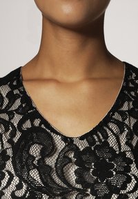 Černý krajkový top s výstřihem do V, zdobený složitými květinovými vzory a průsvitnou texturou. Materiál vypadá lehký a jemný.