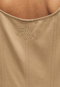 Μπεζ ύφασμα με κάθετες ραφές, διακοσμημένο με κεντημένο κείμενο σε καλλιγραφική γραφή "Hugo" κοντά στο κέντρο πάνω.