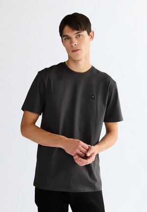 Ung mand med kort mørkt hår iført en ensfarvet mørkegrå T-shirt med korte ærmer, stående mod en lys baggrund, hænderne løst foldet.