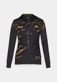 Svart zip-up jakke med gull accenter, hette og teksturert tigerstripe mønster. "UFC" logoen er tydelig vist på brystet.