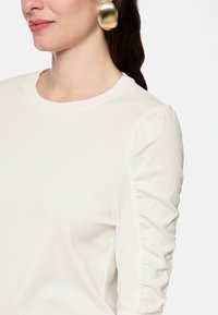 White long-sleeve top with a round neckline and ruched detailing on the sleeves. The fabric is smooth, showcasing a minimalist design.
