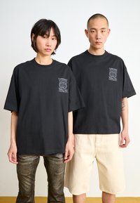 Black oversized t-shirts made of cotton, featuring a pocket graphic design. One model wears dark metallic pants, the other beige shorts.