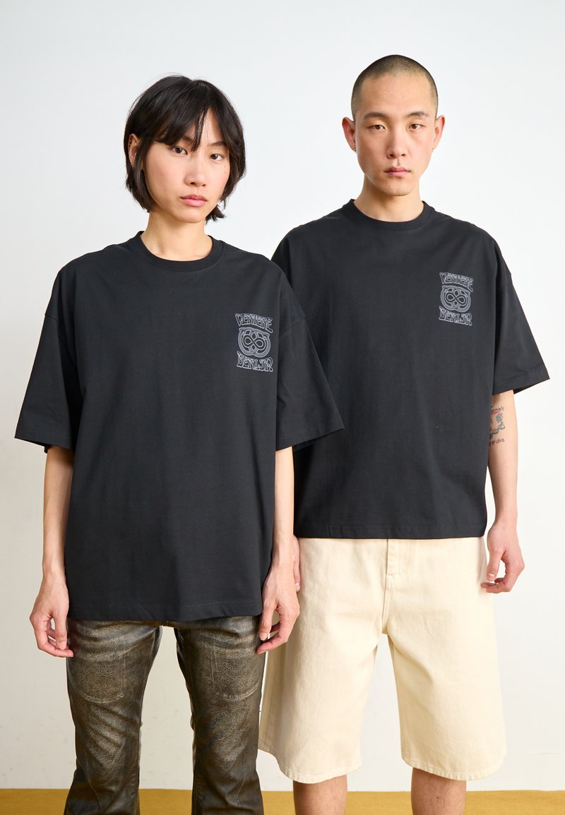 Black oversized t-shirts made of cotton, featuring a pocket graphic design. One model wears dark metallic pants, the other beige shorts.