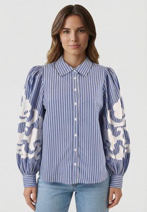 Femme portant un chemisier boutonné à rayures verticales bleues et blanches avec des manches bouffantes ornées de broderies florales blanches, associé à un jean bleu clair.