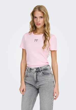 Woman with long blonde hair wearing a light pink short-sleeve top with a small bow design, paired with high-waisted gray jeans.