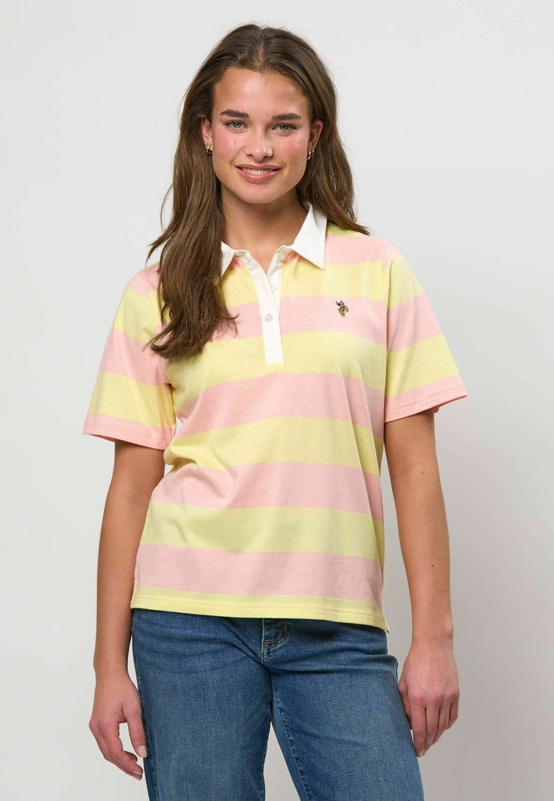 Jeune femme souriante, portant un polo à manches courtes à rayures horizontales rose et jaune, et un jean bleu, debout devant un fond uni.