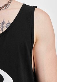 Čierny top bez rukávov vyrobený z bavlny, ktorý má hlboký strih otvorov pre ruky. Detailný záber zobrazuje hladkú textúru a výrazný grafický detail na látke.