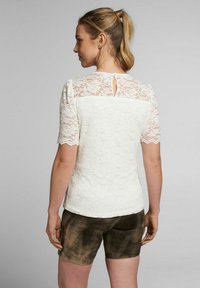 Weiße Spitzenbluse mit kurzen Ärmeln, floralem Muster und einem kleinen Schlüssellochdetail im Rücken. Kombiniert mit braunen Wildleder-Shorts.