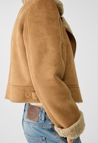 Brązowa zamszowa kurtka z kołnierzem z baranka. Krótki fason, widoczne przeszycia i boczna kieszeń. Noszona na jasnoniebieskich dżinsach z metką.