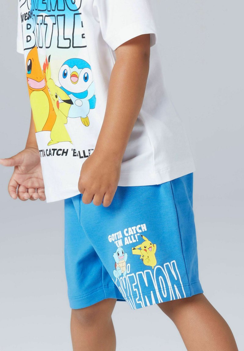Enfant portant un t-shirt blanc et un short bleu avec les personnages Pokémon Pikachu, Salamèche, Tiplouf et Carapuce avec le texte « Gotta catch 'em all ! ».
