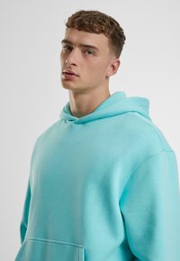 Tirkizna hoodie s opuštenim krojem, s velikim prednjim džepom i mekanom, teksturiranom tkaninom. Minimalistički dizajn s običnom pozadinom.