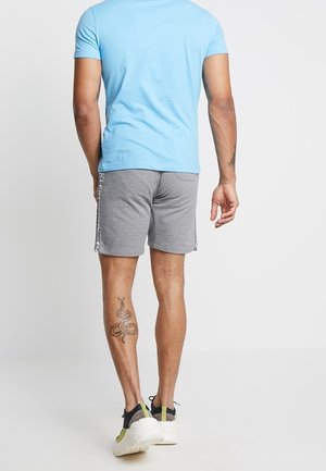 Pantalones cortos deportivos grises con franjas laterales, combinados con una camiseta ajustada azul clara. Llevados con zapatillas deportivas negras y amarillas. Tatuaje de serpiente visible.