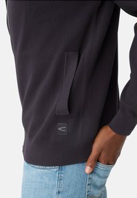 Sort struktureret hættetrøje med sømdetalje, sidelomme og logo-patch. Sammen med lyseblå jeans.