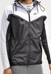 Svart och vit vindtät jacka med dragkedja fram, snörning i huvan, sidofickor och Nike-logotyp på bröstet; har en färgblockdesign.