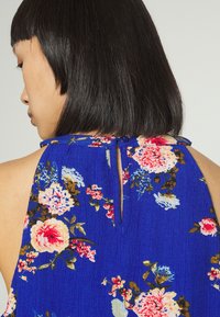 Top floral azul com tecido texturizado, apresentando um detalhe de abertura posterior em forma de chave e fecho com botão. Flores em rosa, branco e verde.
