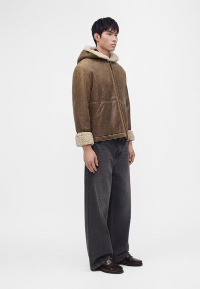 Man standing wearing brown shearling jacket with hood, wide dark gray pants, and dark brown shoes against plain gray background.