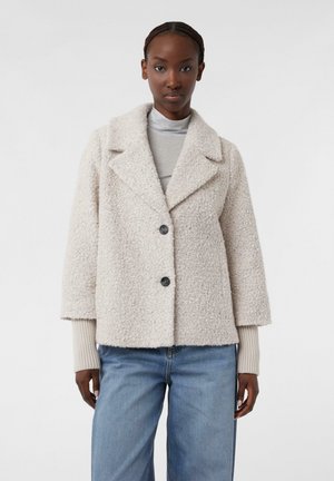 Giacca beige testurizzata con colletto a scansonatura, due grandi bottoni neri e maniche a tre quarti. Abbinata a jeans azzurri chiari.
