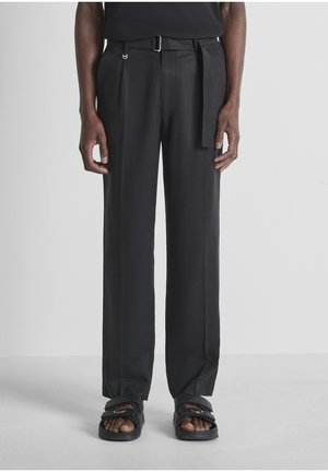 Mannelijk model draagt zwarte broek met wijde pijpen en riem, gecombineerd met zwarte sandalen met open tenen, staand tegen een effen lichte achtergrond.