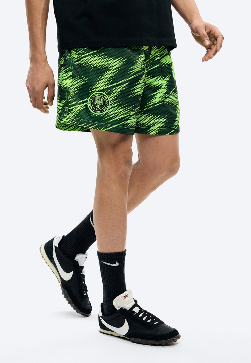 Άτομο που φοράει σορτς με πράσινο και μαύρο μοτίβο και λογότυπο της Ομοσπονδίας Ποδοσφαίρου της Νιγηρίας, μαύρες κάλτσες Nike και μαύρα αθλητικά παπούτσια Nike.