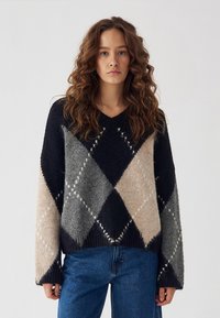 Femeie cu păr lung și creț purtând un pulover cu model argyle în negru, gri și bej și blugi albaștri, pe un fundal simplu.