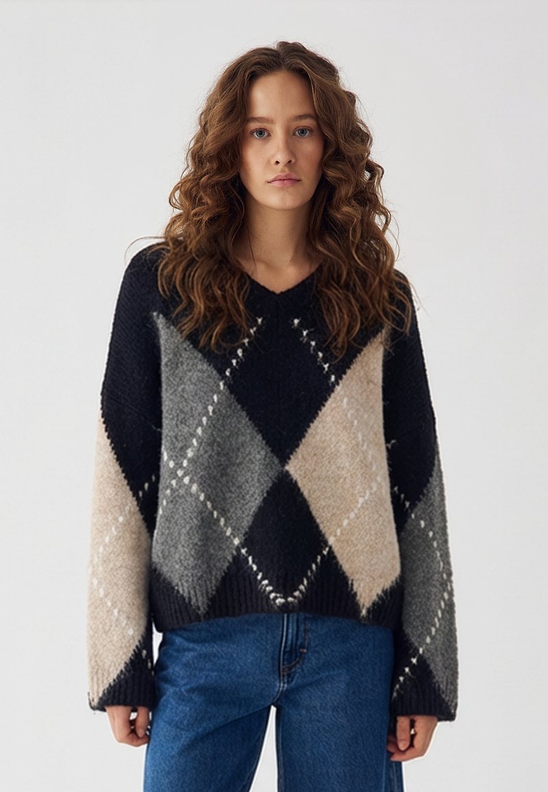 Femeie cu păr lung și creț purtând un pulover cu model argyle în negru, gri și bej și blugi albaștri, pe un fundal simplu.