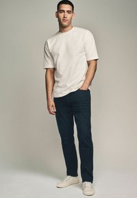 Tricou din bumbac crem, taiere lejeră, mâneci scurte, asortat cu pantaloni navy închis și adidași de culoare deschisă, stand împotriva unui fundal neutru.