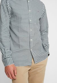 Camisa de cuadros verde y blanca de botones con mangas largas, de tela ligera y con dobladillo curvado. Presenta un sutil logo en el pecho.