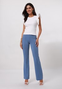 Haut blanc à côtes avec des manches volantées, associé à un pantalon bleu évasé à la texture lisse et aux détails de coutures. Porté avec des sandales blanches.