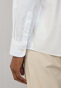 Camicia bianca a maniche lunghe con polsini a bottone, caratterizzata da una texture liscia e una vestibilità su misura. Esibita su una mano su uno sfondo neutro.