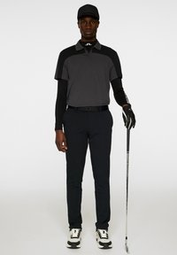 Svart golfoutfit bestående av en kortärmad polo med grå detaljer, långa ärmar under, svarta byxor och svart keps. Håller en golfklubba.