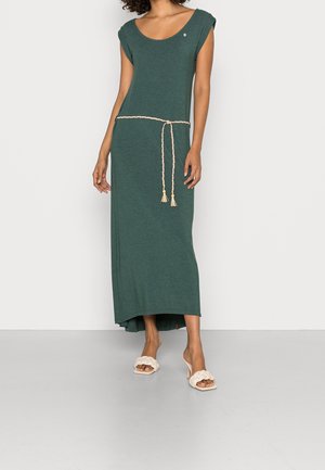 Kobieta ubrana w ciemnozieloną, krótkorękawową sukienkę sięgającą do kostek, z plecionym beżowym paskiem i białymi sandałami na obcasie.