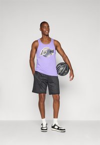 Un débardeur violet avec le logo des Los Angeles Lakers, associé à un short en maille noir. Tenant un ballon de basket, portant des baskets noires et blanches.
