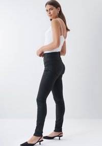 Zwarte high-waisted broek van een gladde stof, met een slanke pasvorm en duidelijke naadlijnen. Gepaard met een witte tanktop en zwarte hakken.