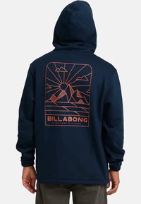 Mężczyzna w granatowej bluzie z kapturem z pomarańczową grafiką góry i słońca oraz napisem "BILLABONG Adventure Division" z tyłu.