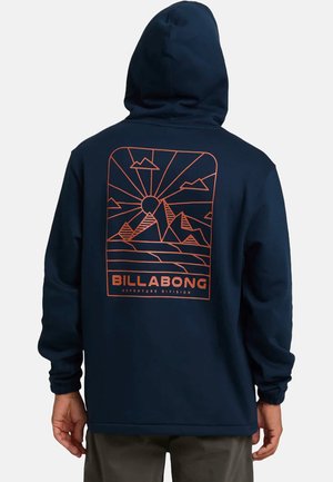 Uomo che indossa una felpa con cappuccio blu navy con una grafica di una montagna arancione e un sole, e il testo "BILLABONG Adventure Division" sulla schiena.
