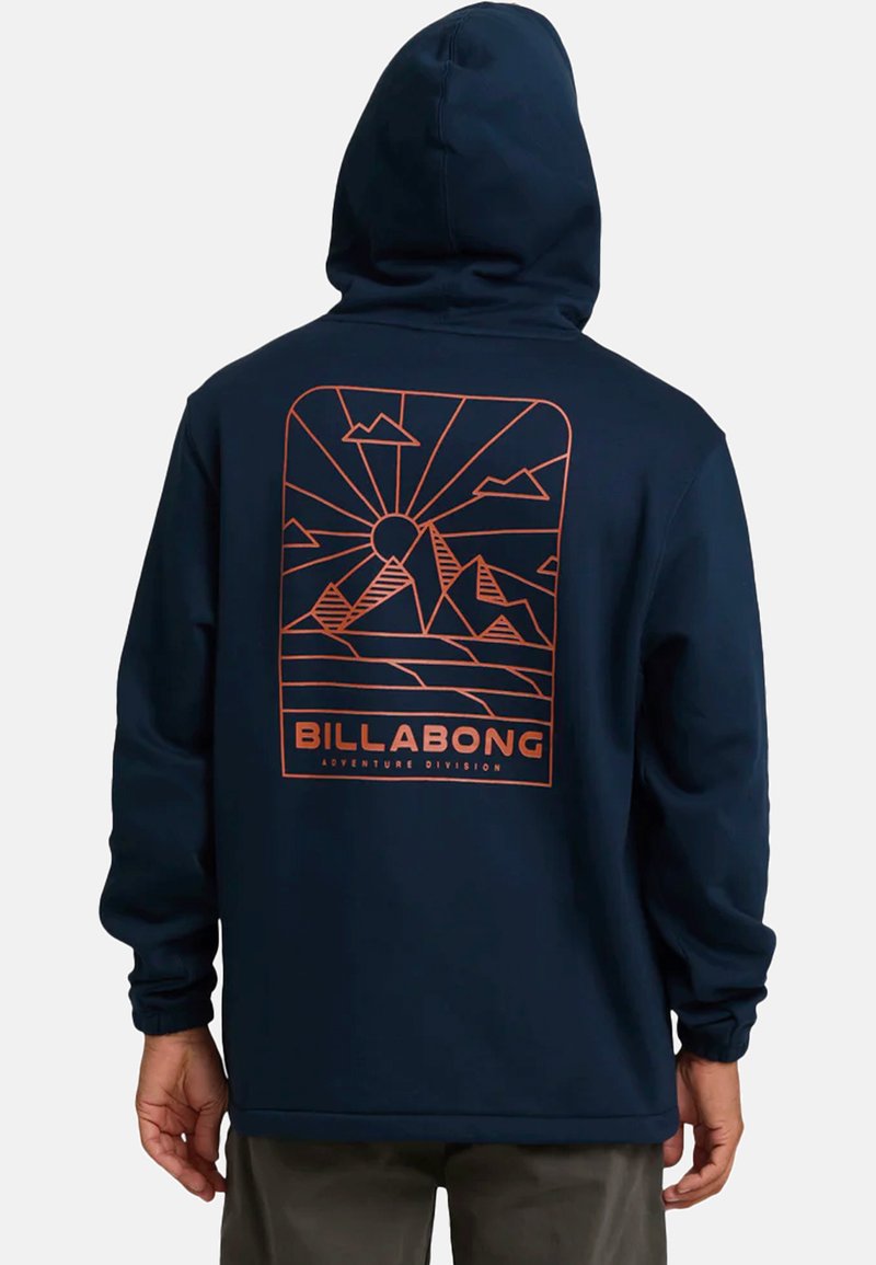 Mężczyzna w granatowej bluzie z kapturem z pomarańczową grafiką góry i słońca oraz napisem "BILLABONG Adventure Division" z tyłu.