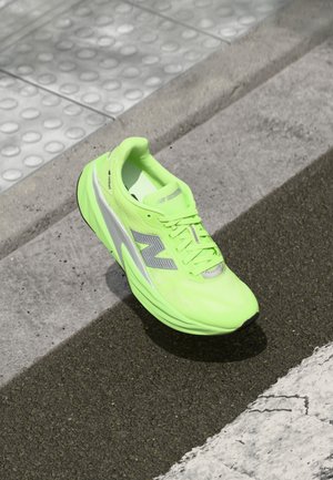 Baskets de course New Balance d'un vert néon lumineux reposant sur un pavé texturé près d'une surface de marche tactile et du bord d'un trottoir.