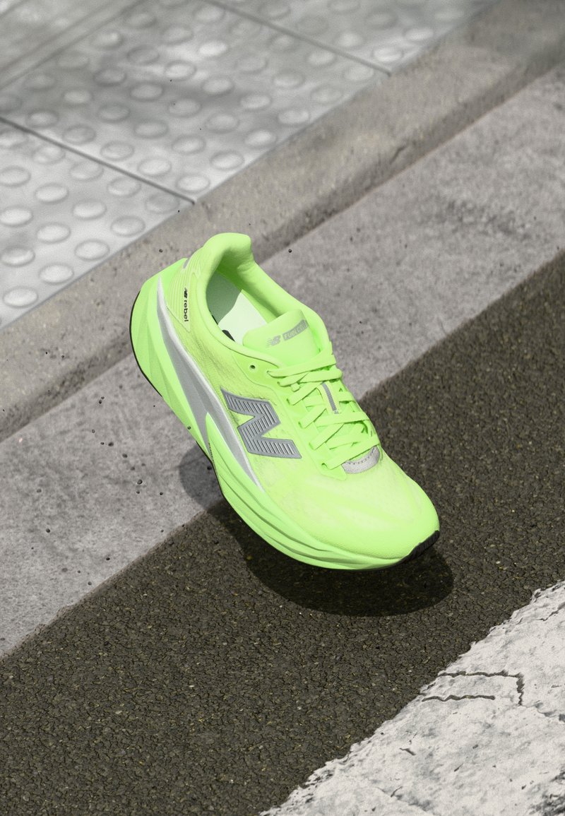 Sjajna neon zelena New Balance tenisica za trčanje leži na teksturiranom asfaltu blizu taktilne površine za hodanje i rubnika.